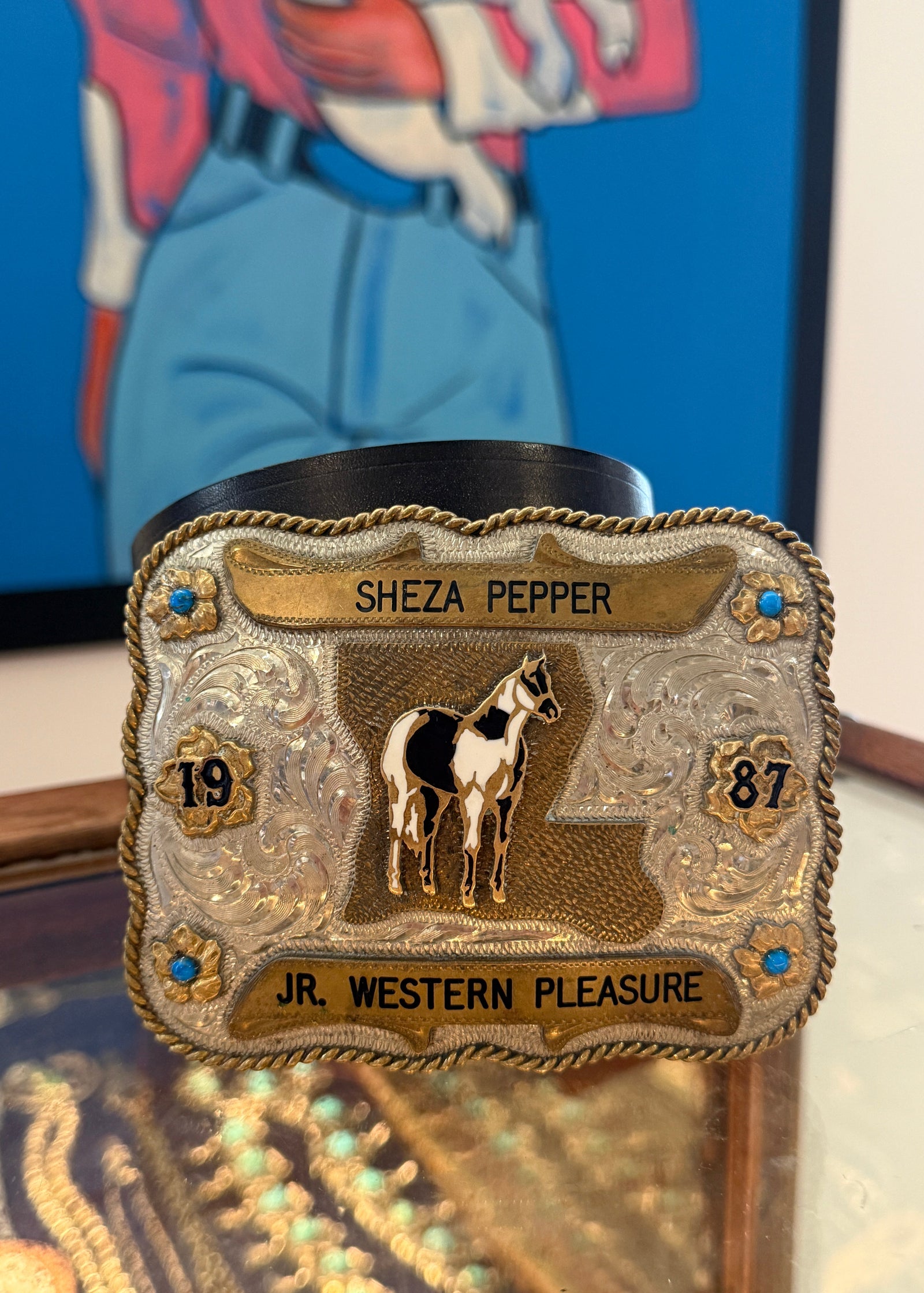 A decorative western-style belt buckle with engraved text reading 'SHEZA PEPPER' and 'JR. WESTERN PLEASURE,' featuring a horse illustration and small blue stone accents, photographed indoors against a colorful framed artwork.