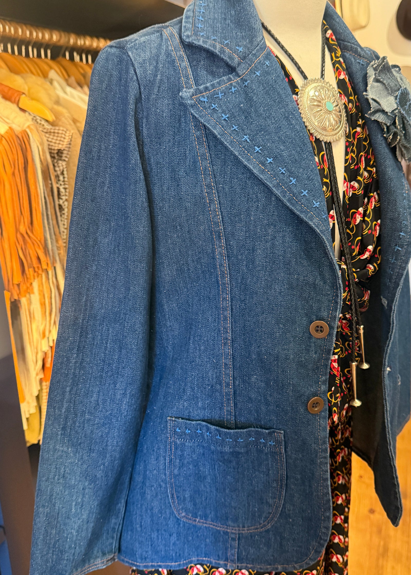 A blue denim blazer displayed on a mannequin torso in a clothing store. The blazer features notched lapels, contrast stitching, a front button closure, and a large front patch pocket.