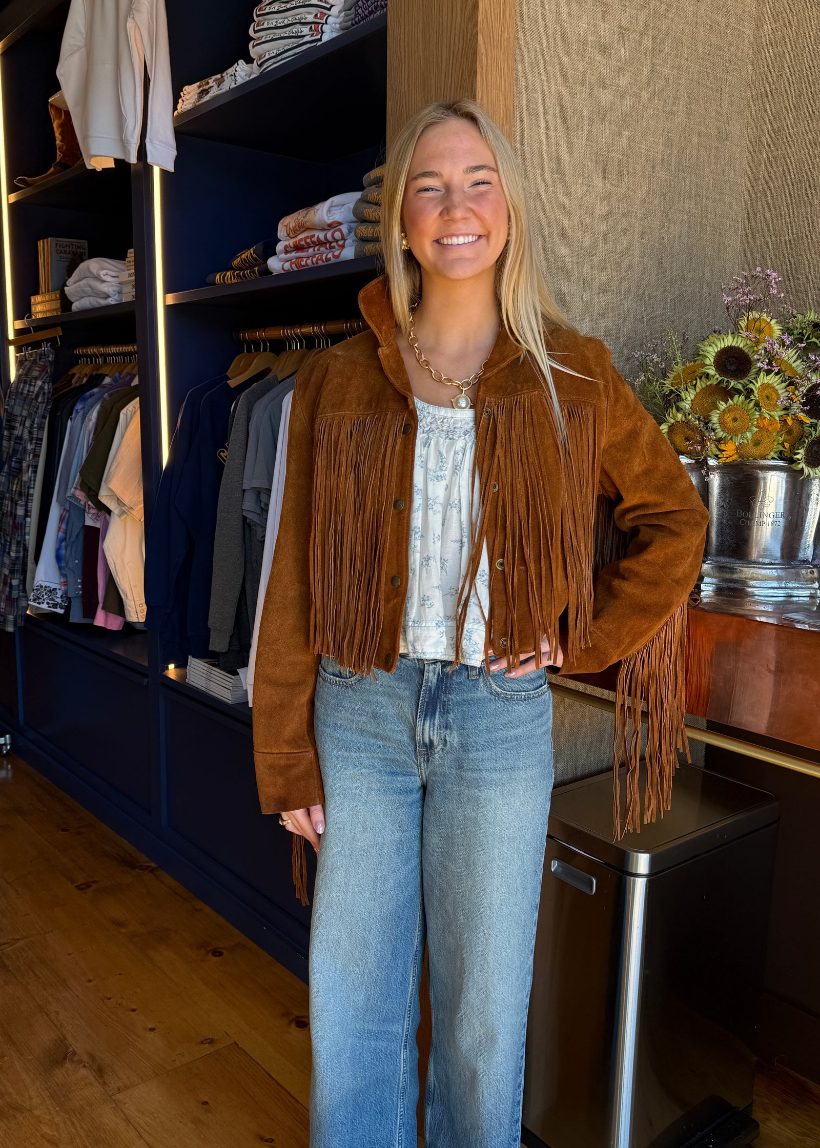 A woman stands in a boutique closet area wearing a brown suede fringe jacket over a white top and blue jeans.