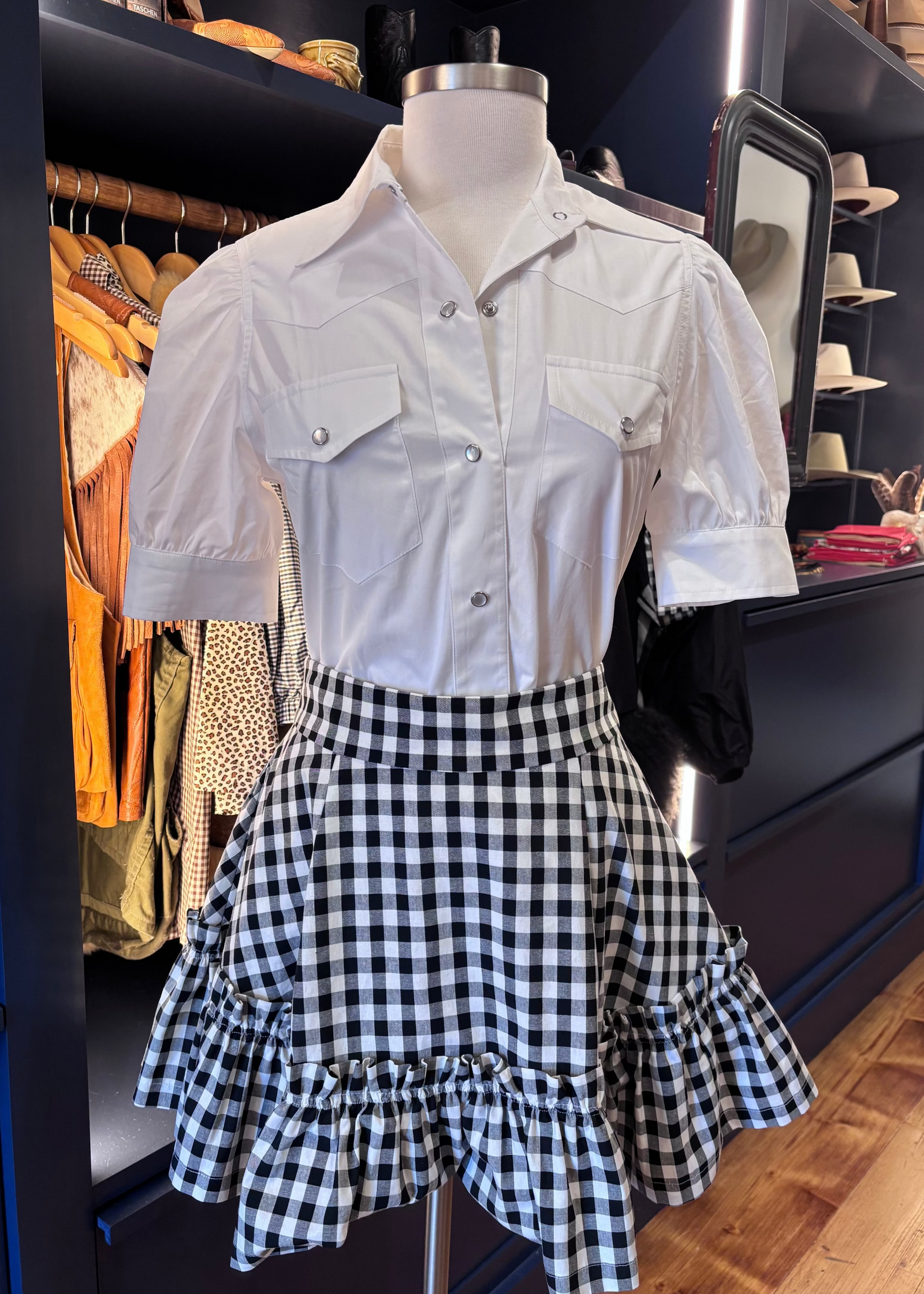 A white short-sleeve button-front shirt with chest flap pockets is paired with a black-and-white gingham ruffled skirt, displayed on a mannequin in a clothing boutique.