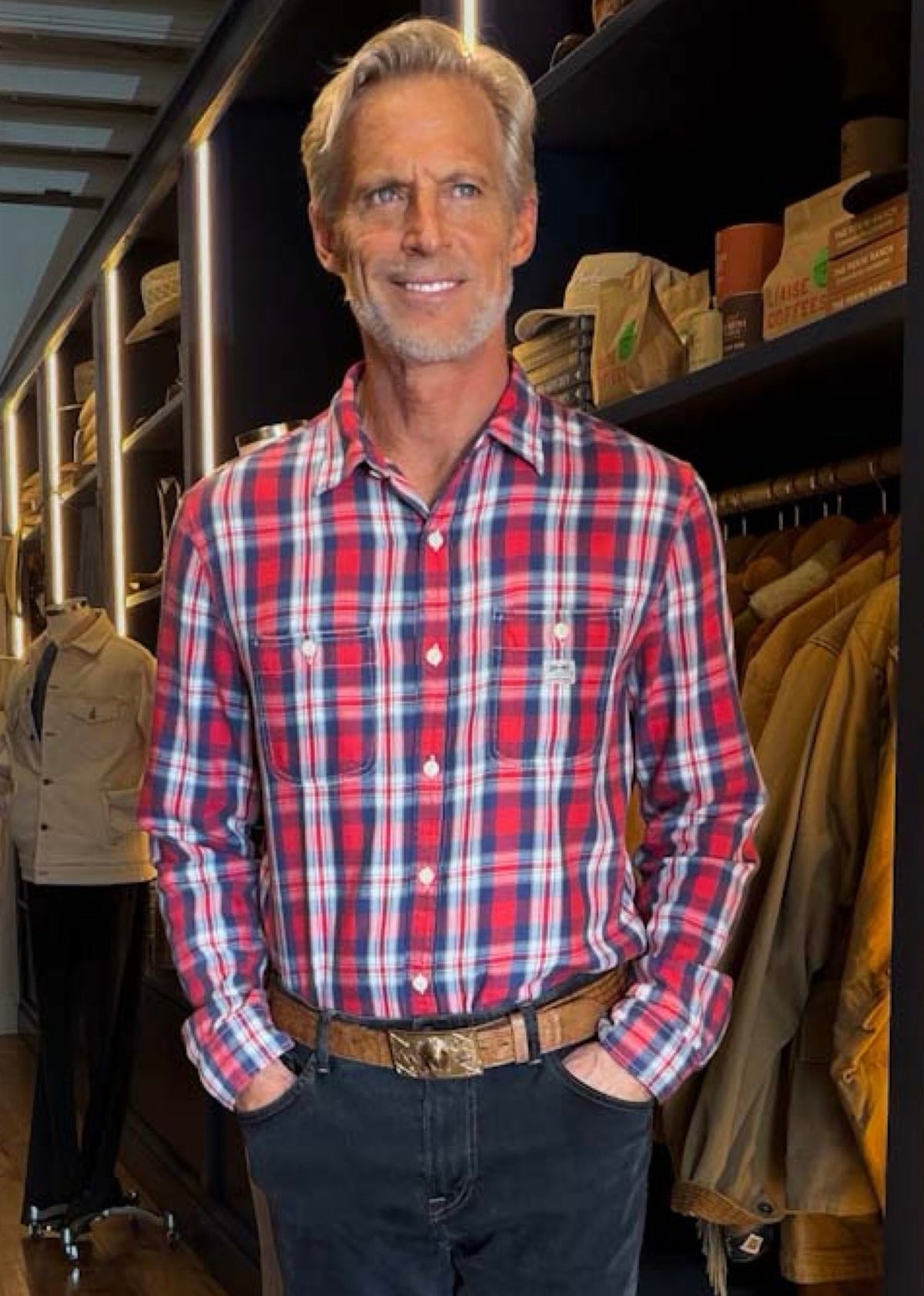 A smiling man with gray hair stands in a retail store wearing a red, white, and blue plaid button-down shirt, dark gray jeans, and a brown leather belt.