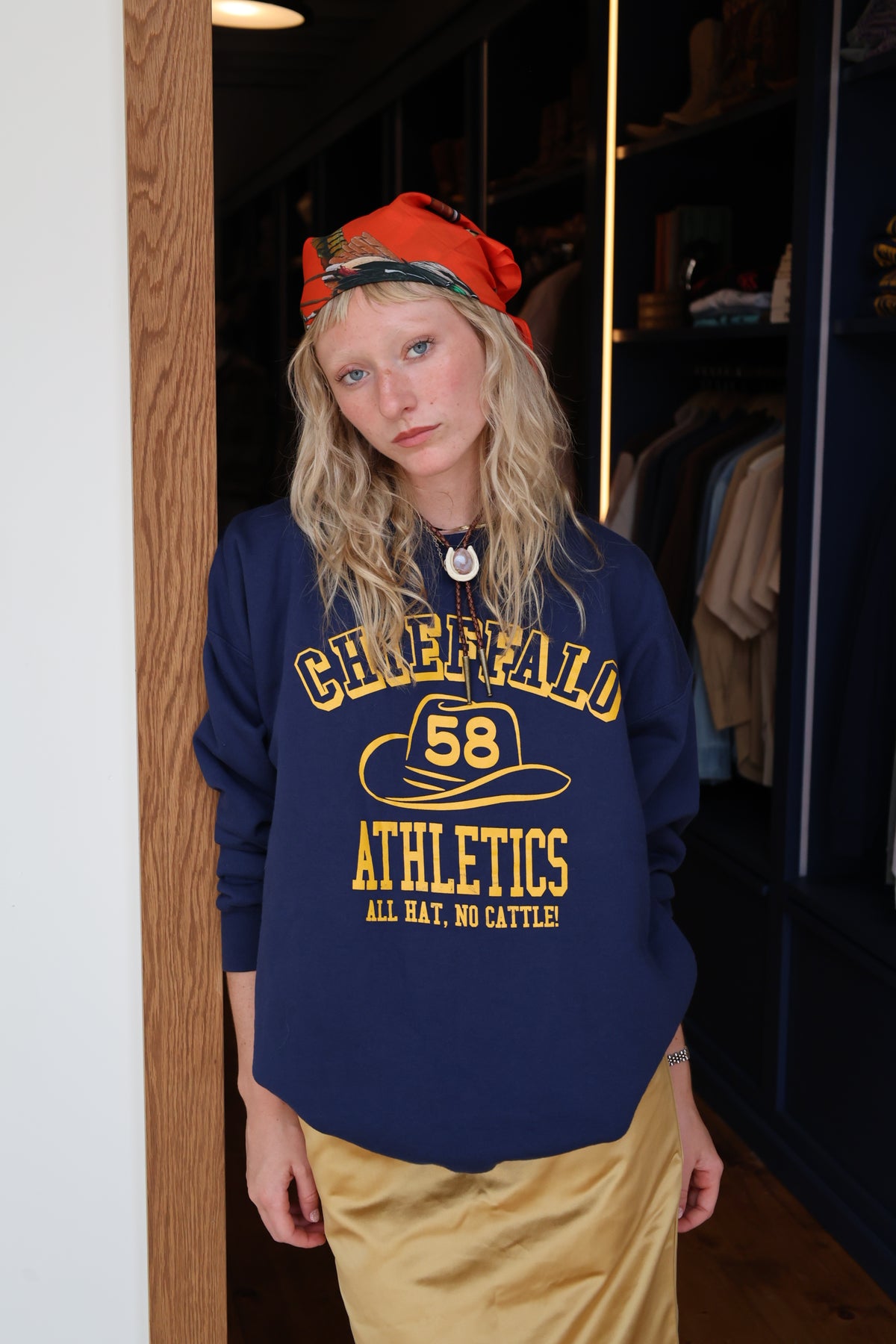 A woman wearing a navy blue sweatshirt with yellow 'CHIEFFALO 58 Athletics' text, paired with a red bandana and tan pants, standing in a closet area.