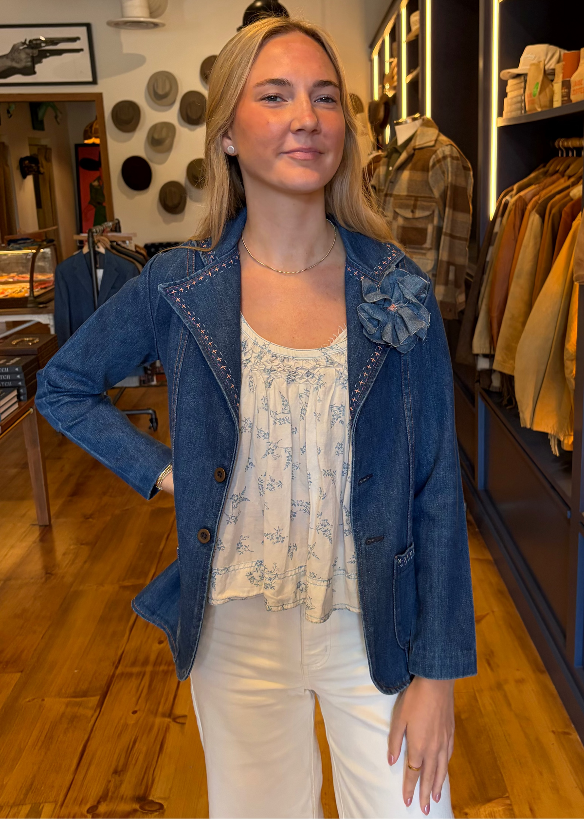 A woman posing in a clothing store wearing a blue denim jacket with decorative stitching and a floral applique, paired with a white floral top and white pants.