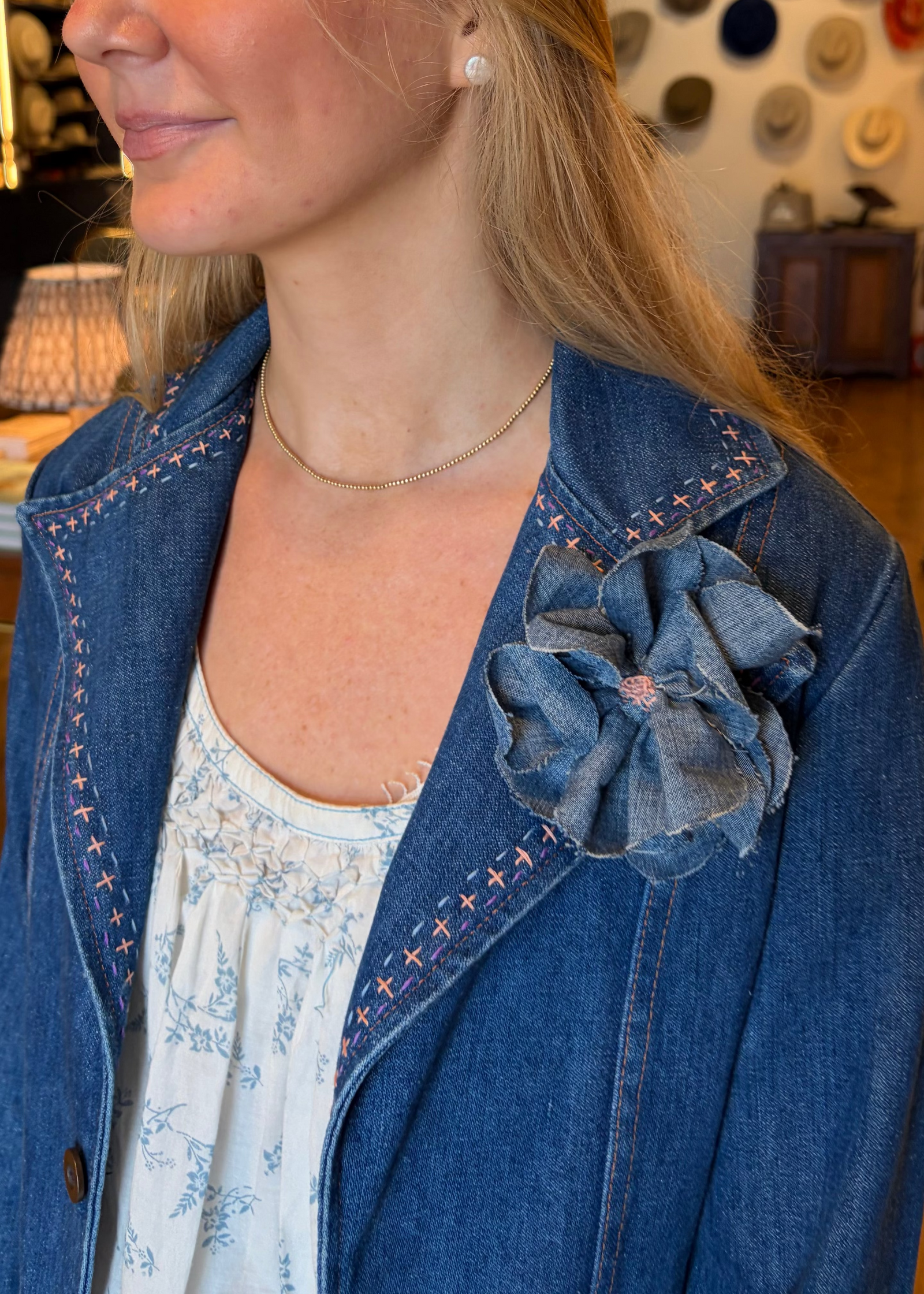 A close-up lifestyle shot of a woman wearing a blue denim jacket with decorative contrast stitching and a matching denim flower accessory pinned to her lapel, over a white patterned top.