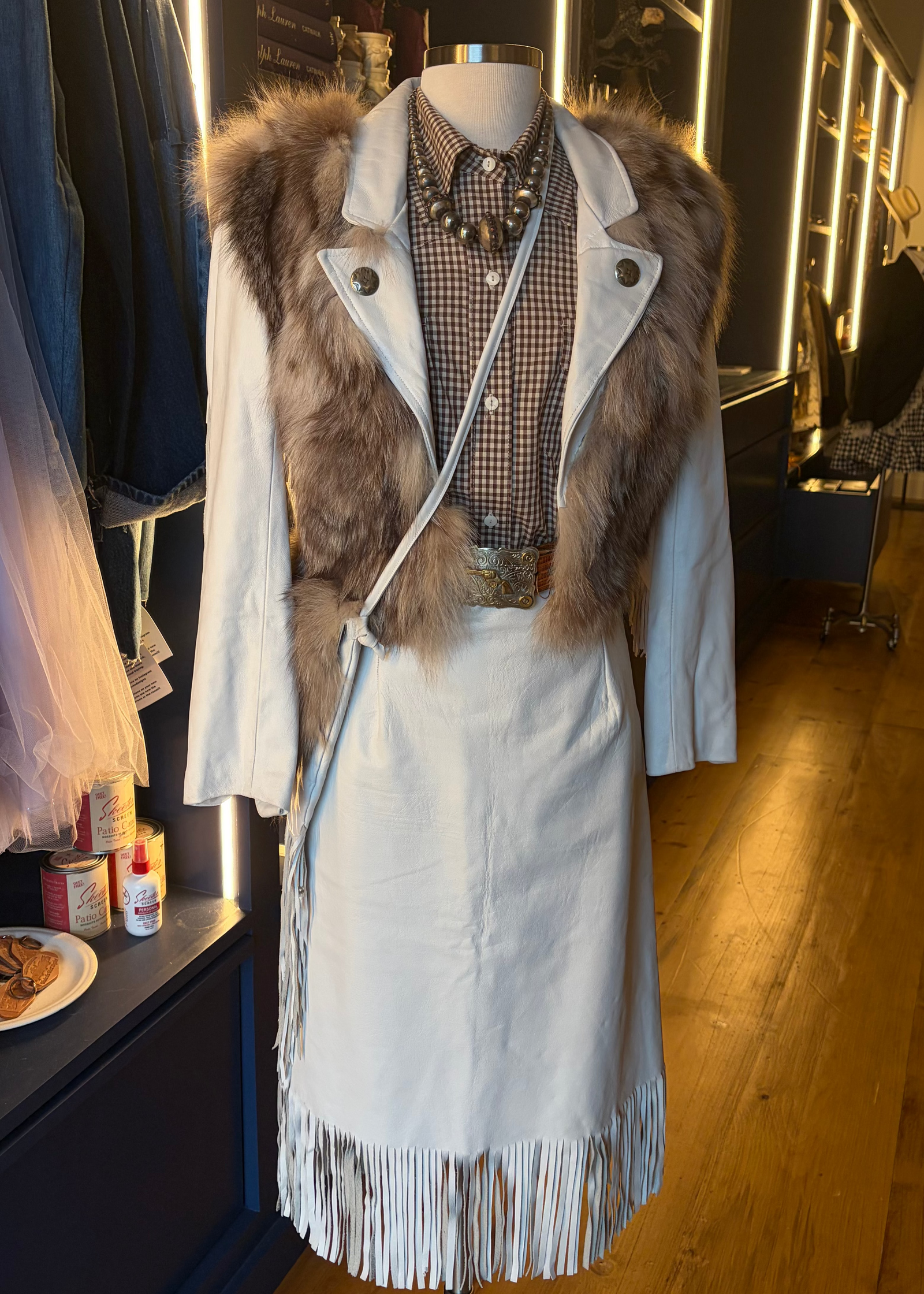 A light blue fringe dress displayed on a mannequin in a clothing boutique, styled with a brown fur-trimmed vest, a patterned shirt, layered necklaces, and a silver belt.
