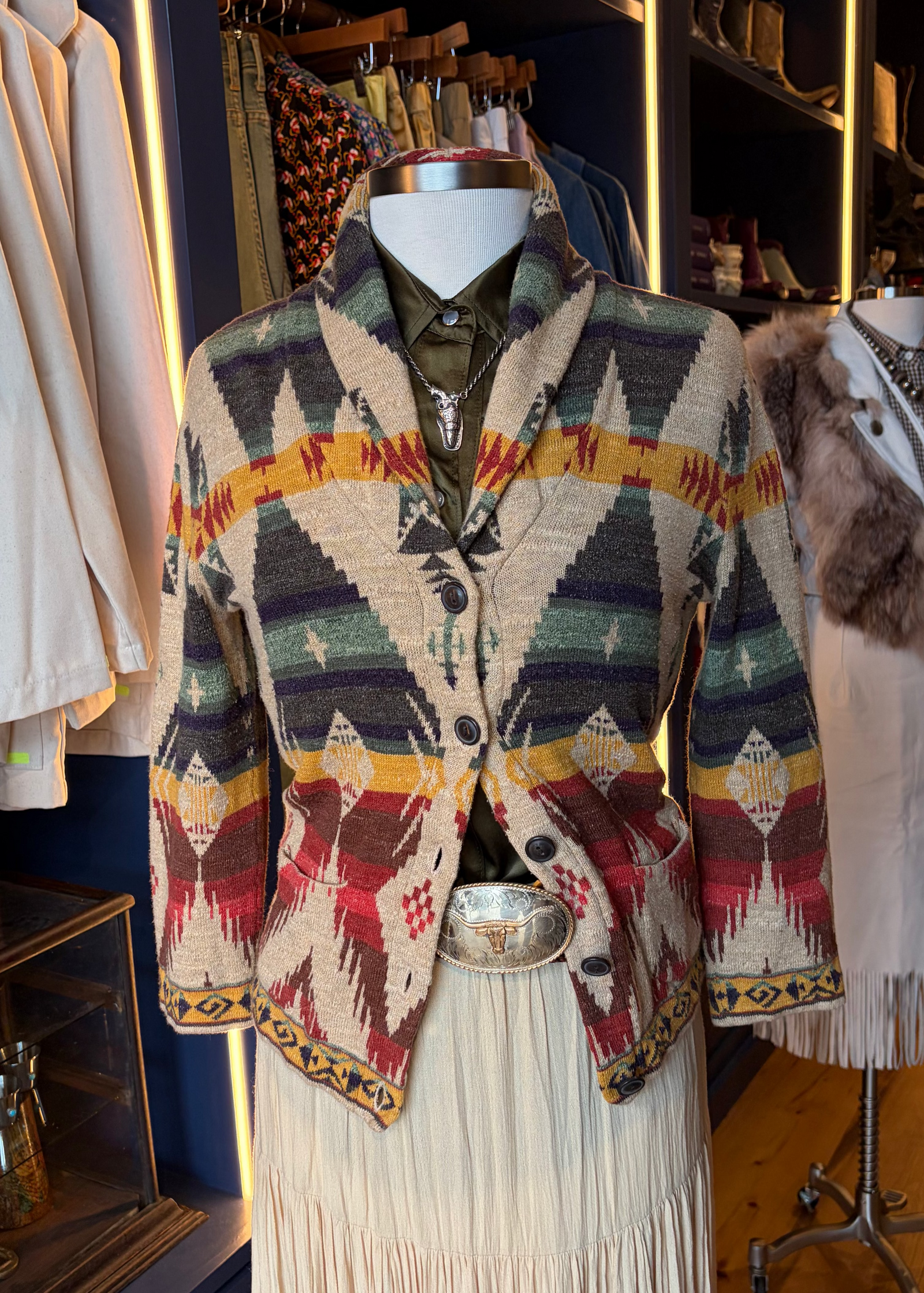A patterned knit cropped cardigan with a geometric Southwestern design in beige, red, green, yellow, and black, styled on a mannequin with a cream pleated skirt and belt in a boutique setting.