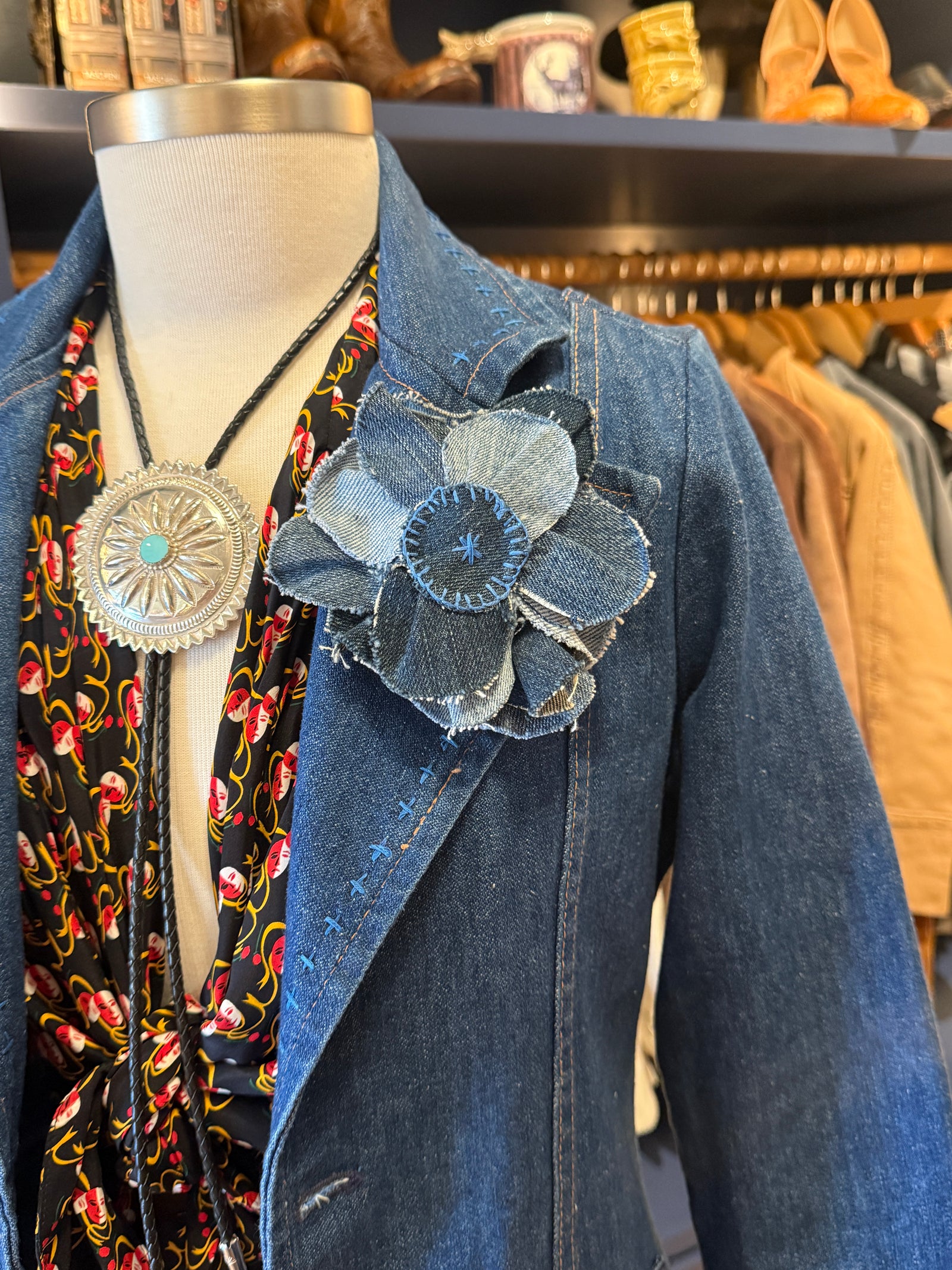 A denim jacket displayed on a mannequin torso, styled with a black floral scarf, a large silver-toned pendant necklace, and a large fabric flower embellishment on the lapel. The background shows a clothing store interior with racks of garments.
