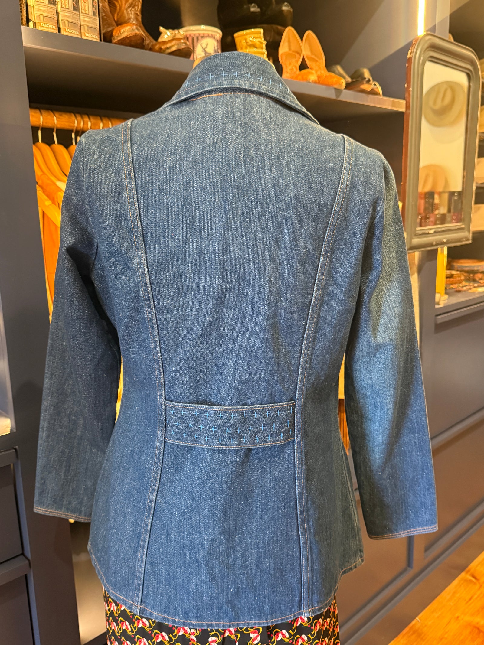 A blue denim blazer jacket displayed on a mannequin from the back, featuring a structured collar, three-quarter sleeves, and a decorative buttoned welt pocket at the waist, set against a boutique interior background.
