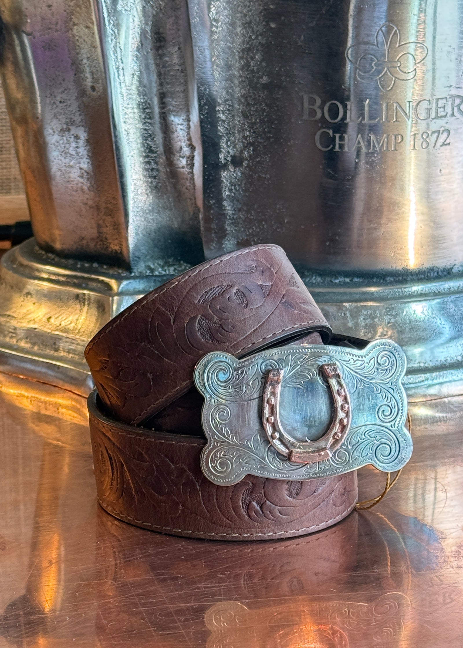 A brown tooled leather belt with a silver horseshoe-shaped buckle, coiled on a reflective copper surface in front of a large silver metal container.