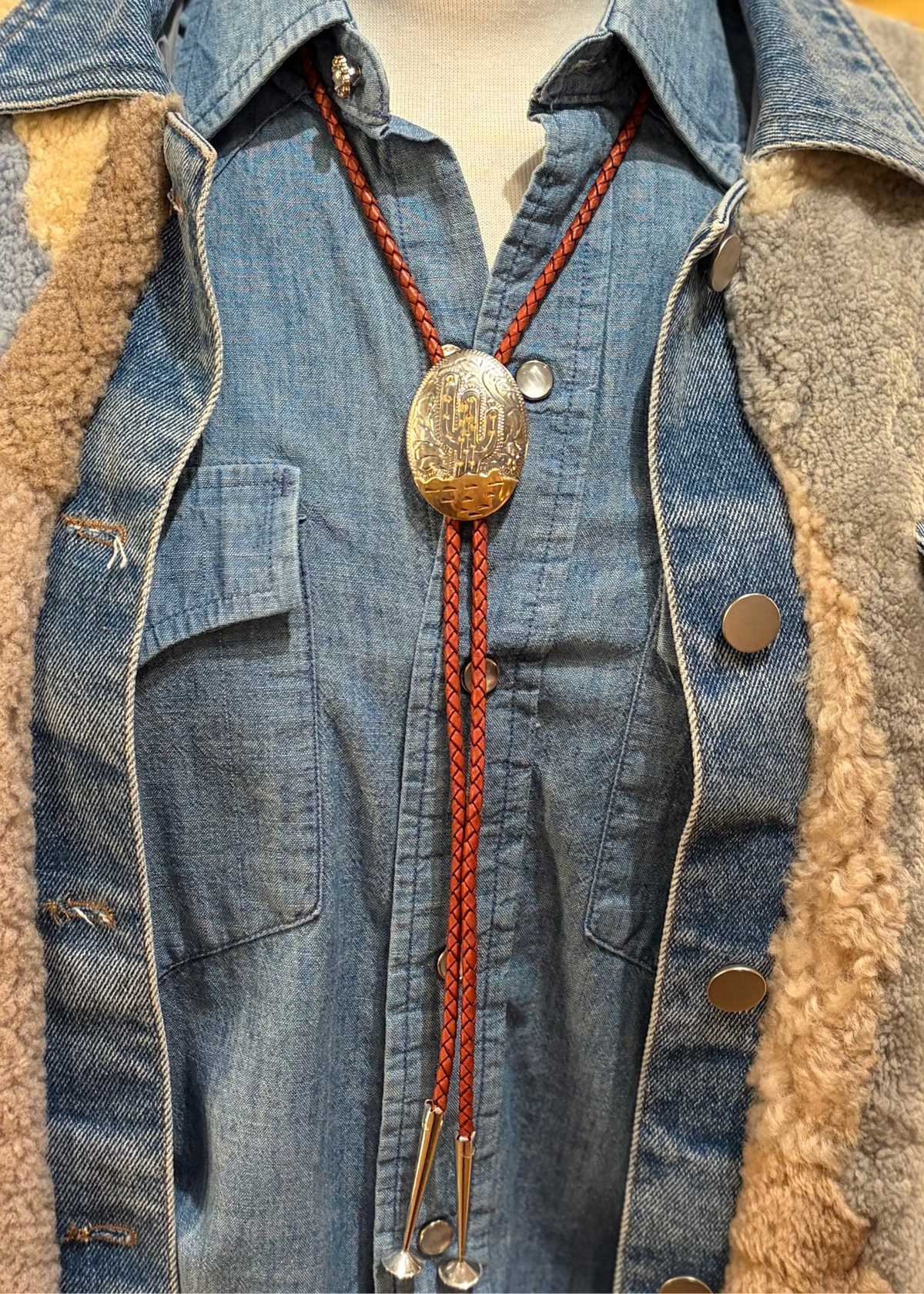 A close-up of a denim shirt jacket with a tan shearling lining, styled with a braided red cord bolo tie featuring a large gold-toned engraved oval medallion.