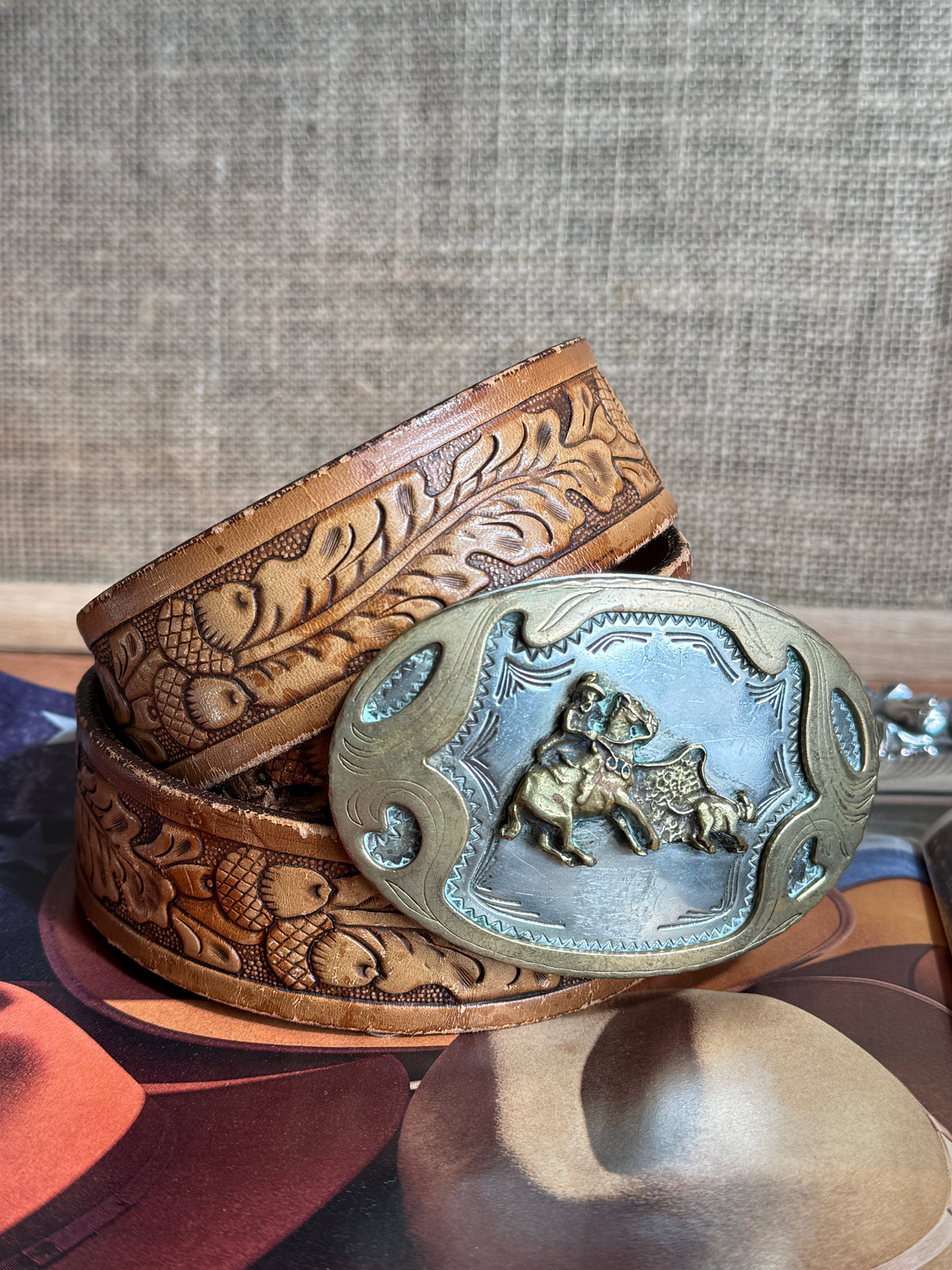 A tooled leather belt with a large oval metal buckle featuring a raised cowboy and steer motif, displayed on a surface against a textured fabric backdrop.