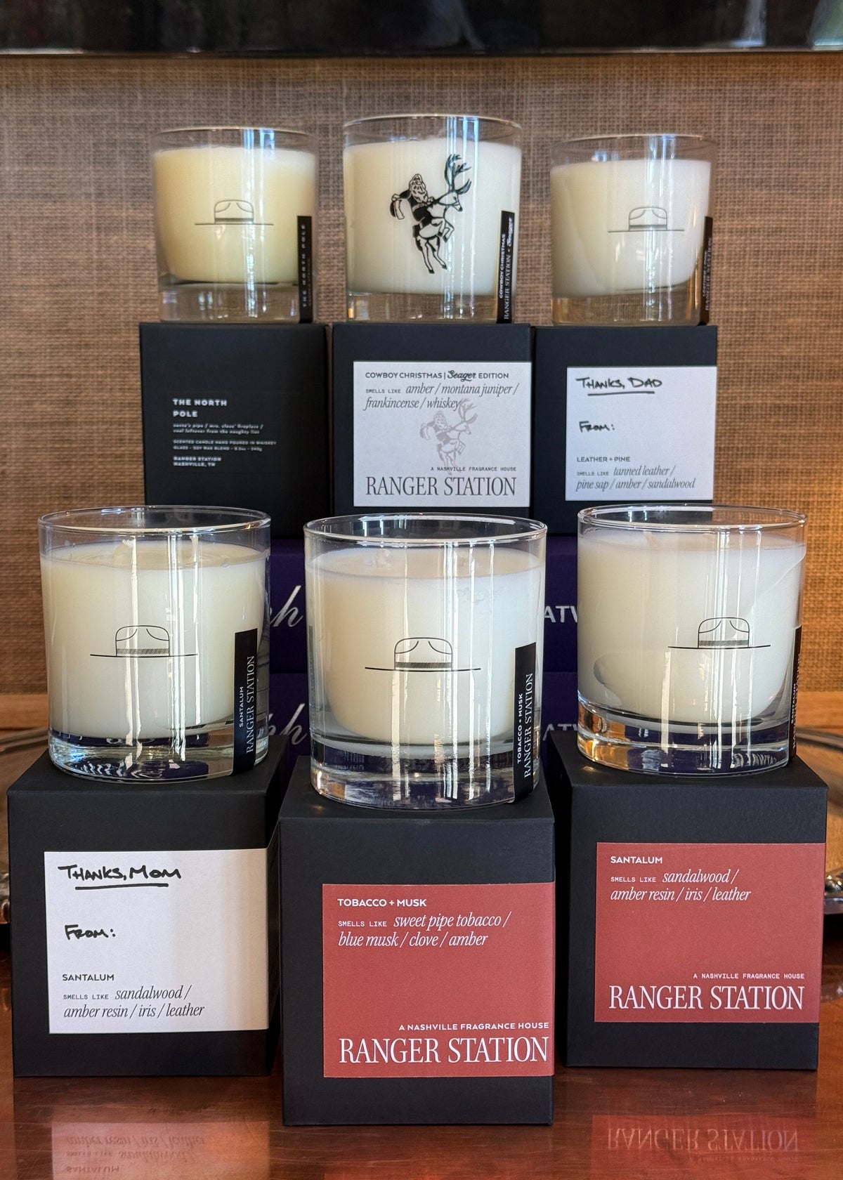 Six scented candles in clear glass jars with cream-colored wax, arranged alongside their black packaging boxes on a brown wooden surface. The boxes feature a reddish-brown label with white text reading 'RANGER STATION'.