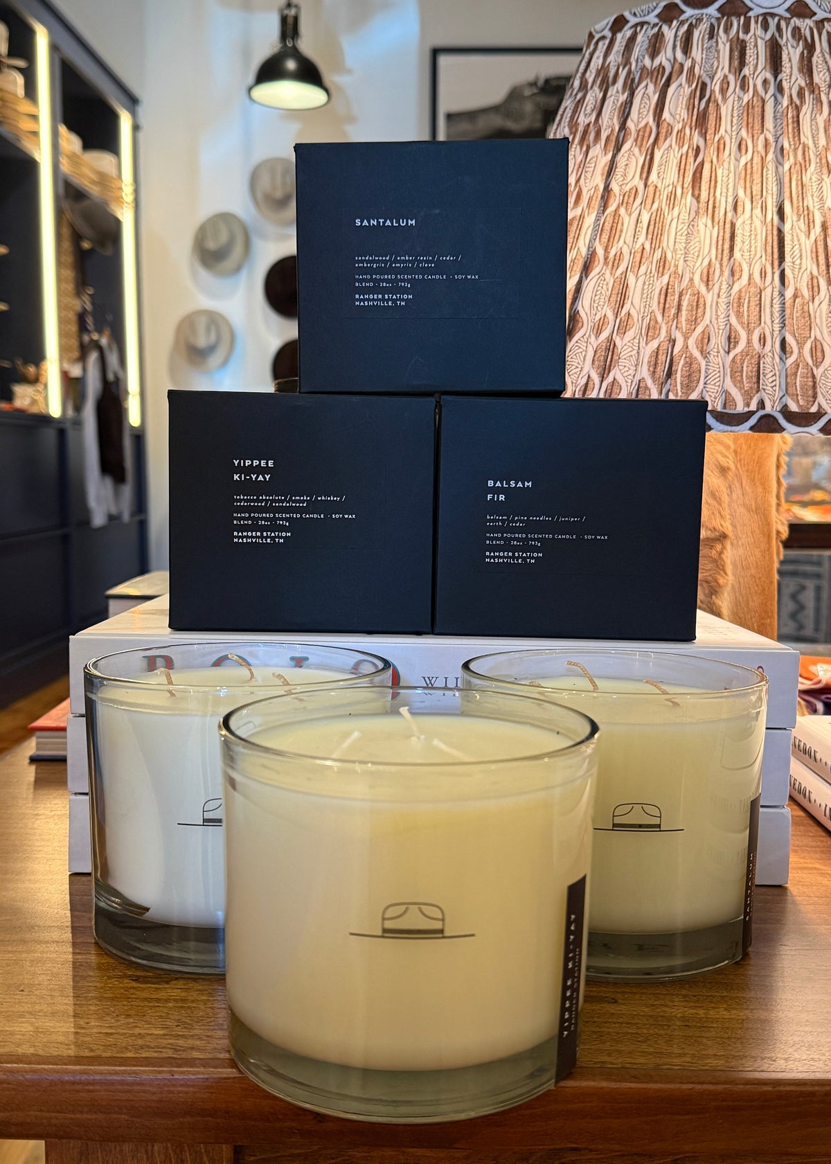 Three Santalum candles in clear glass jars with white wax are displayed on a wooden table, with three matching black product boxes stacked behind them in a boutique setting.