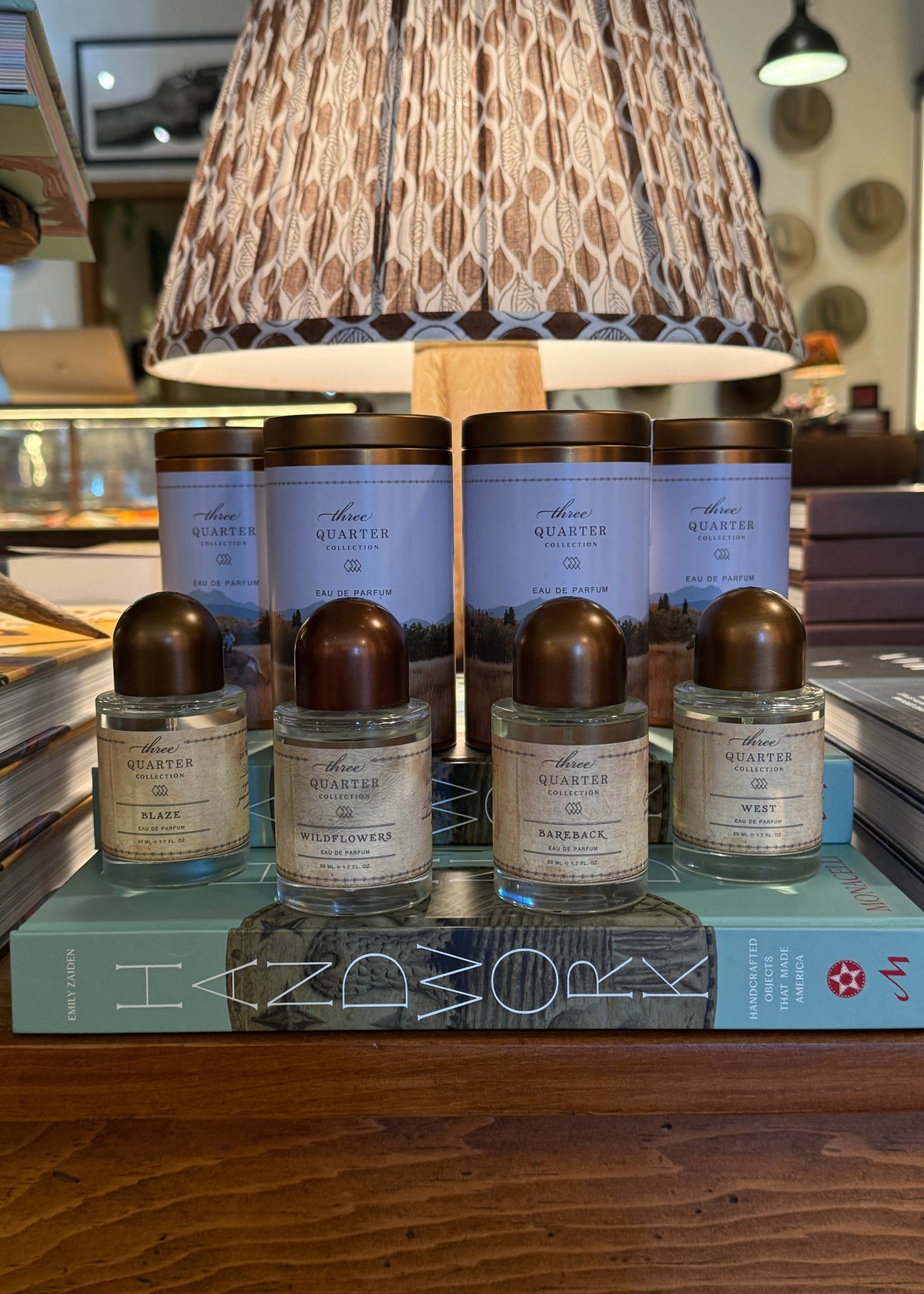 A set of Quatered home fragrance products displayed on a wooden shelf in a retail setting, featuring four glass bottles with metallic bronze caps and four cylindrical jars with light blue labels and bronze lids, arranged on top of a teal book in front of a patterned lampshade.