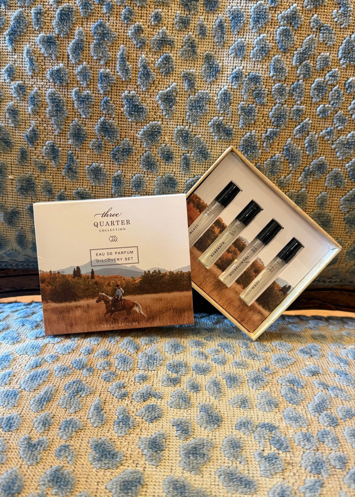 A boxed perfume gift set labeled 'Three Quarter Scenic Ride' with one closed box and one open box showing four small perfume vials, placed on a blue-and-beige patterned fabric surface.